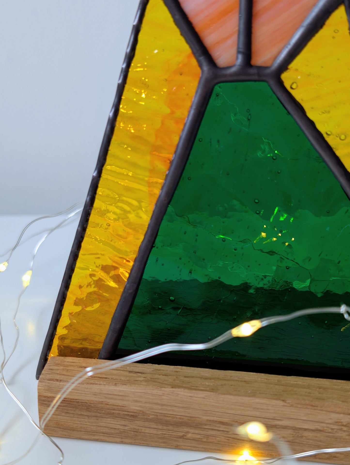 Triangular stained glass panel with wooden base and string lights on a white background